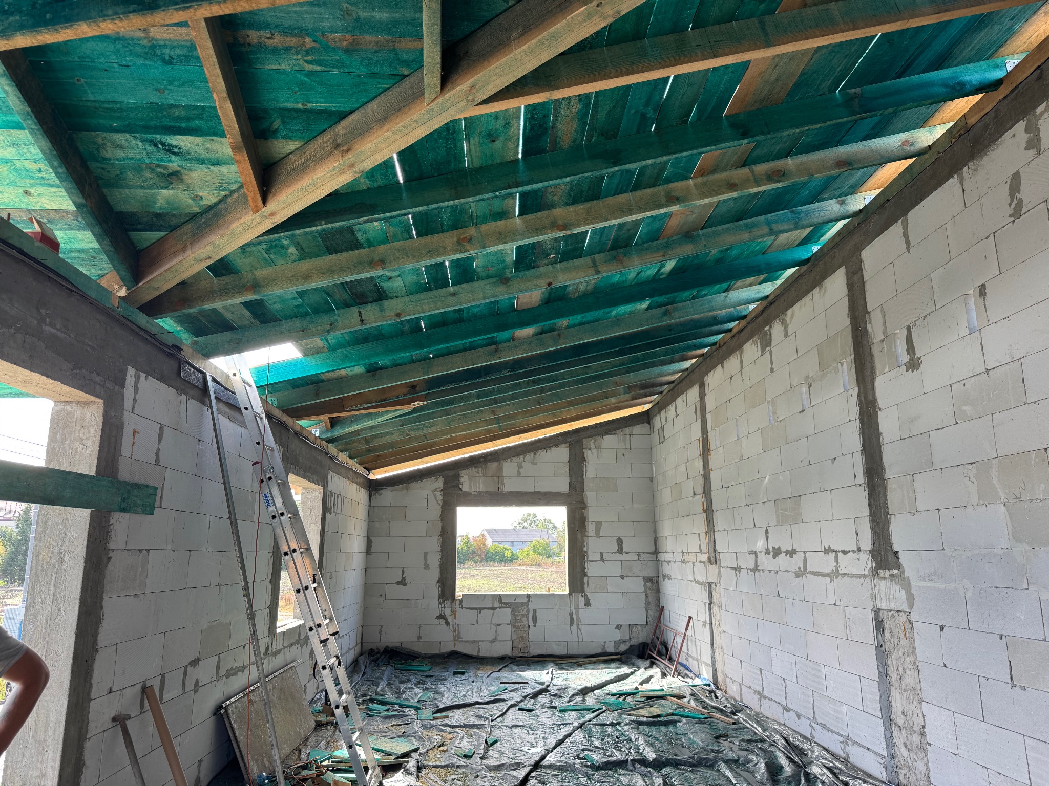 Surowy stan budynku z białych bloczków, widoczne drewniane elementy konstrukcji dachu pomalowane na zielono, drabina oparta o ścianę, okno z widokiem na zewnątrz.