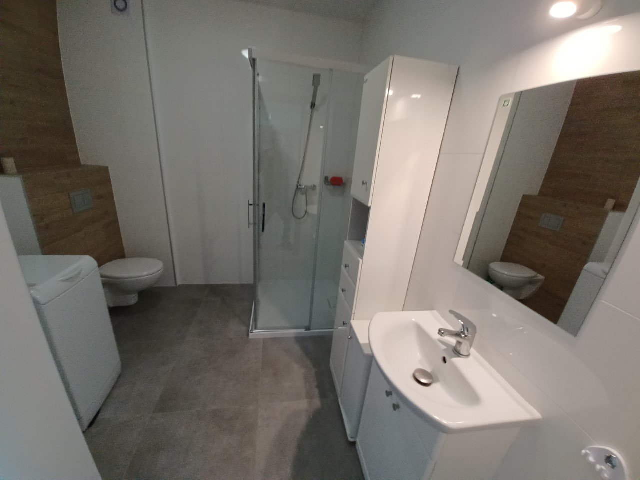 Nowoczesna łazienka z kabiną prysznicową, umywalką nablatową i pralką, wykończona w bieli i szarości z akcentem drewnopodobnej okładziny ściennej.