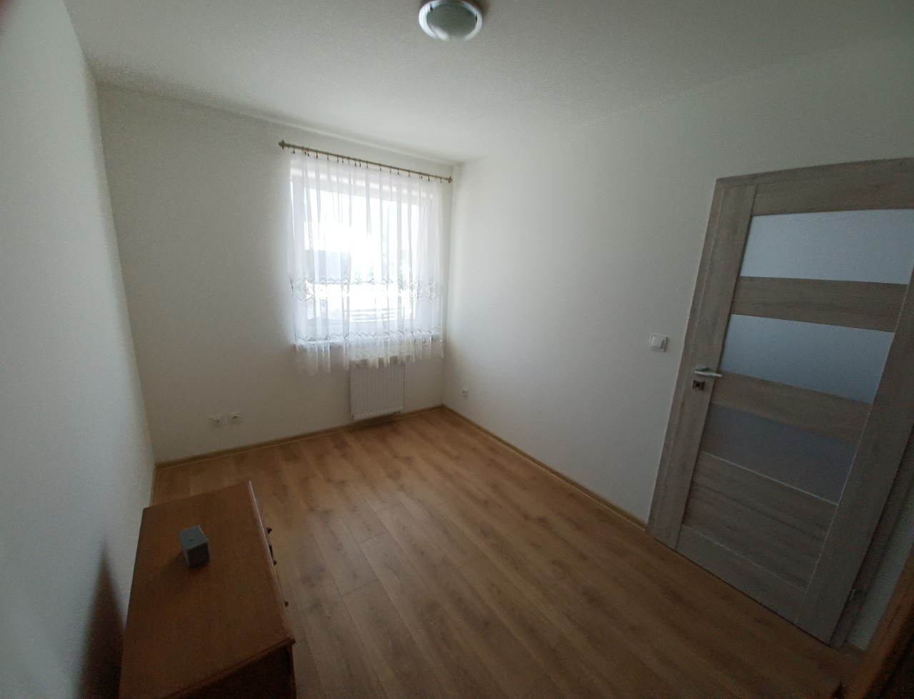 Puste, jasne pomieszczenie z drewnianą podłogą, oknem z białymi firankami i grzejnikiem pod nim, oraz drewnianymi drzwiami z przeszkleniami. Na podłodze stoi komoda z kostką na blacie.