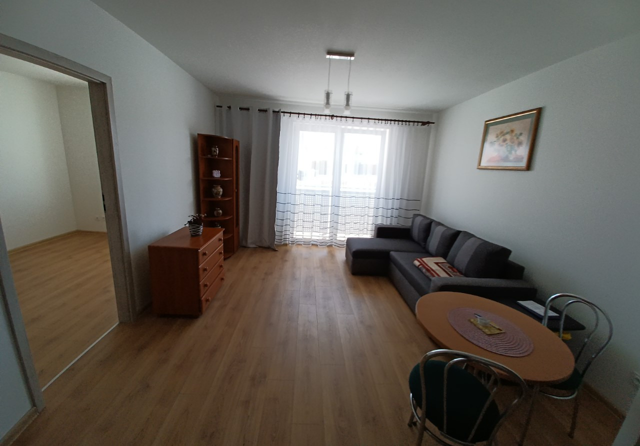 Jasne wnętrze salonu z drewnianą podłogą, szarą sofą narożną, okrągłym stołem z dwoma krzesłami, komodą i regałem, oświetlone naturalnym światłem wpadającym przez okno z białymi zasłonami.