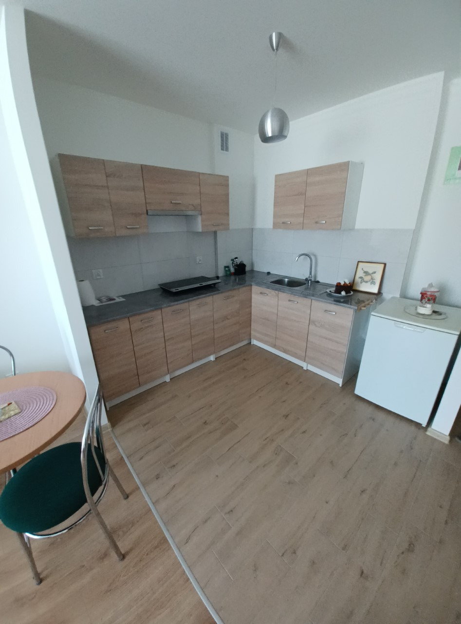 Jasna kuchnia z drewnianymi szafkami, szarym blatem, okrągłym stołem z zielonym krzesłem i małą lodówką, oświetlona nowoczesną lampą wiszącą.