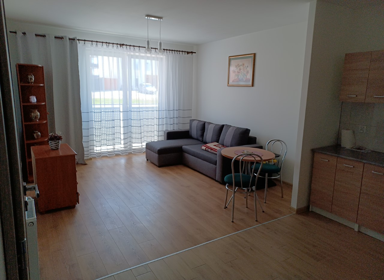 Jasny salon z rozkładaną szarą sofą, okrągłym stolikiem z dwoma krzesłami, drewnianą komodą i regałem, widok na balkon przez firanki.