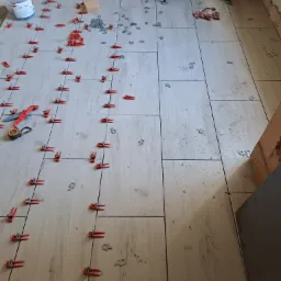 Układanie jasnych płytek podłogowych w łazience, widoczne narzędzia i system poziomowania płytek, wanna na nóżkach w tle.