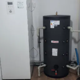 Biała pompa ciepła Panasonic Aquarea i czarny zbiornik Huch EnTEC z podłączeniami hydraulicznymi na tle białej ściany i brązowej podłogi imitującej drewno.