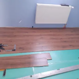 Rozpoczęty montaż paneli podłogowych na zielonej piance izolacyjnej, widoczne narzędzia: kątownik, nóż, poziomica oraz dystanse, ściana pomalowana na jasny niebieski kolor, grzejnik na ścianie.