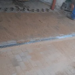 Montaż liniowego odwodnienia na brukowanym podłożu, zasypane piaskiem, widoczne fragmenty kostki brukowej i robocze ubranie