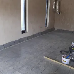 Układanie szarych płytek podłogowych w surowym wnętrzu, widoczne narzędzia: poziomica, odkurzacz przemysłowy i wiadro z zaprawą.