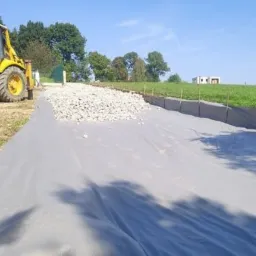 Żółta koparko-ładowarka Komatsu obok przygotowanego podłoża z geowłókniny i warstwą kruszywa, widok na teren budowy z elementami krajobrazu wiejskiego.