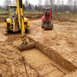 FIRMA USŁUGOWA PAWEŁ FOKSIŃSKI - Żółta koparka w trakcie wykonywania wykopu pod fundamenty, widoczna równa krawędź wykopu, w tle czerwona minikoparka i zarys drzew na horyzoncie.