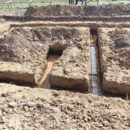 Wykopane rowy pod fundamenty z widocznym zbrojeniem i częściowo wypełnione wodą, ziemia wokół wykopu.