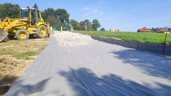 Żółta koparko-ładowarka Komatsu obok przygotowanego podłoża z geowłókniny i warstwą kruszywa, widok na teren budowy z elementami krajobrazu wiejskiego.