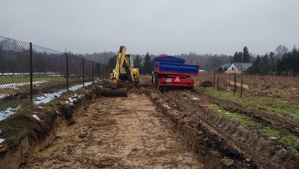 Żółta koparka i niebiesko-czerwona przyczepa na błotnistym placu budowy, widoczny wykop wzdłuż ogrodzenia z siatki, zimowy krajobraz w tle.