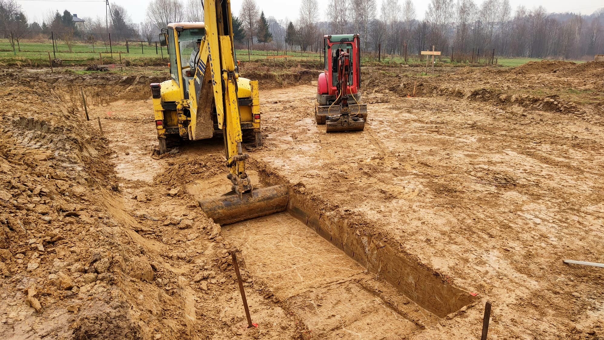 Żółta koparka w trakcie wykonywania wykopu pod fundamenty, widoczna równa krawędź wykopu, w tle czerwona minikoparka i zarys drzew na horyzoncie.