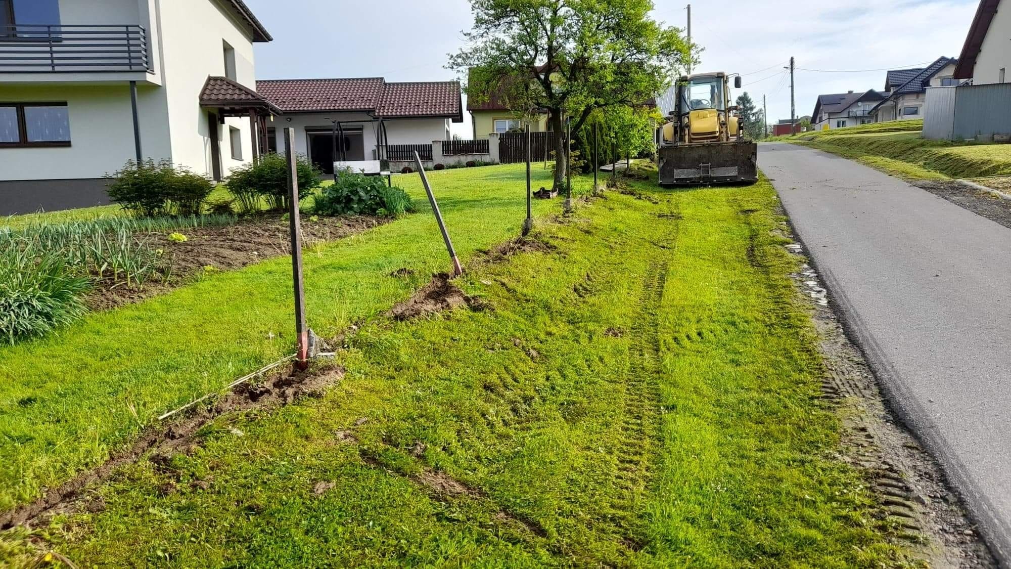 Żółta koparko-ładowarka na trawniku, wytyczona linia palików, ślady opon na trawie, domy w tle.