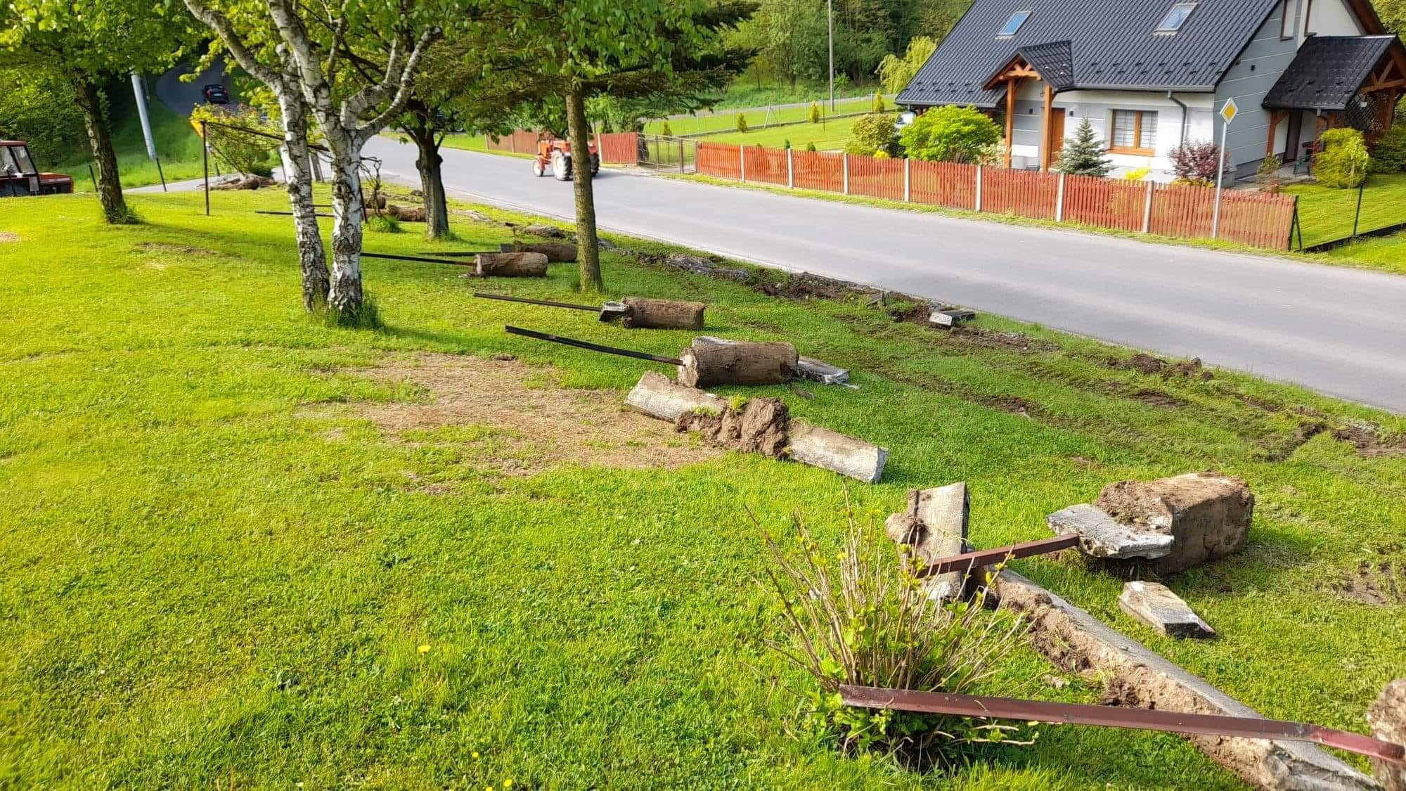 Przygotowanie terenu pod budowę: usunięte fragmenty betonowego ogrodzenia z wystającymi metalowymi elementami leżą na trawie wzdłuż drogi, w tle dom jednorodzinny i traktor.