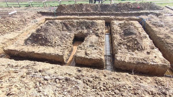 Wykopane rowy pod fundamenty z widocznym zbrojeniem i częściowo wypełnione wodą, ziemia wokół wykopu.