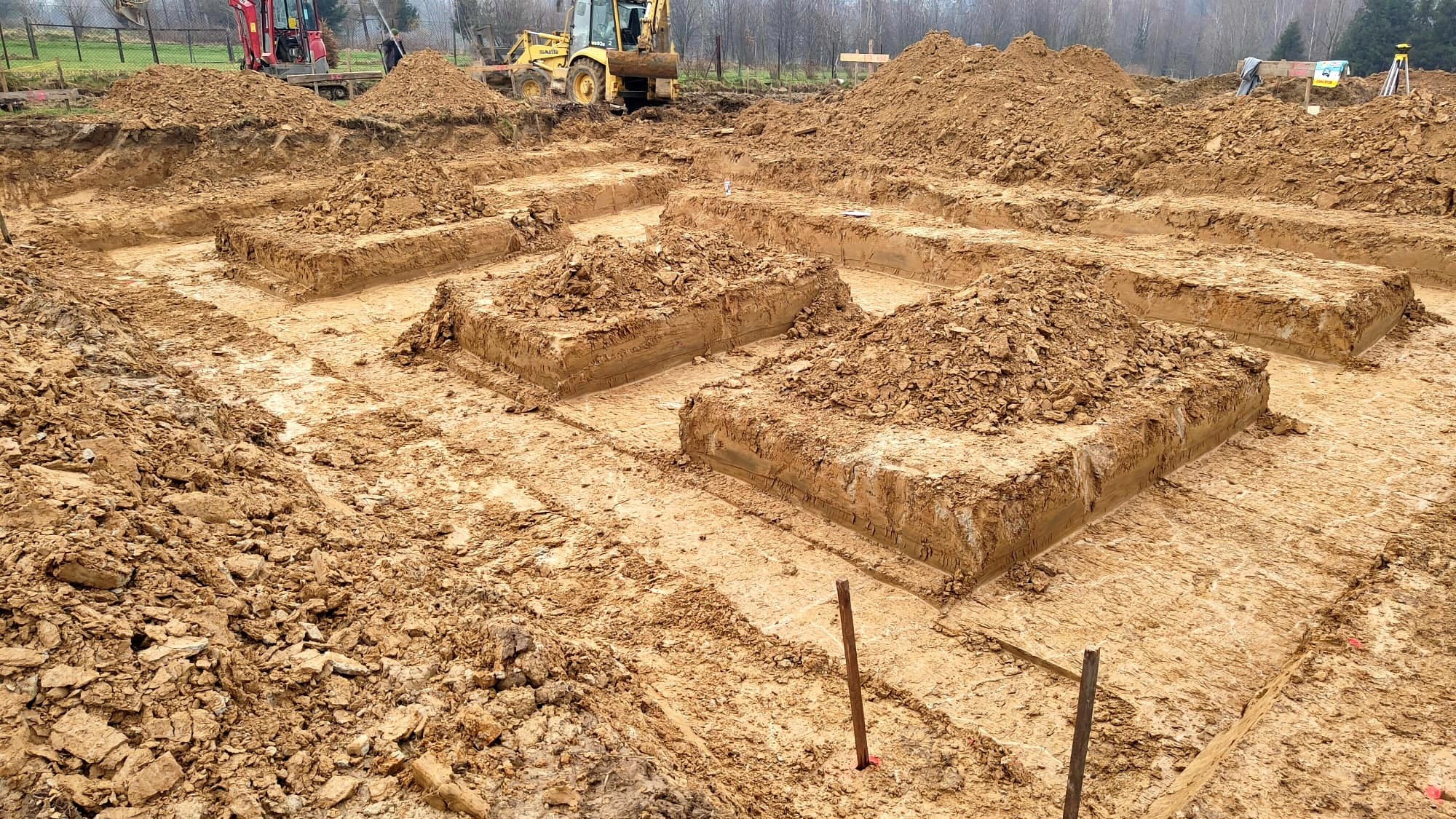 Przygotowanie terenu pod budowę: wykopane rowy pod fundamenty, pryzmy ziemi, widoczny sprzęt budowlany w tle, poziomica laserowa.