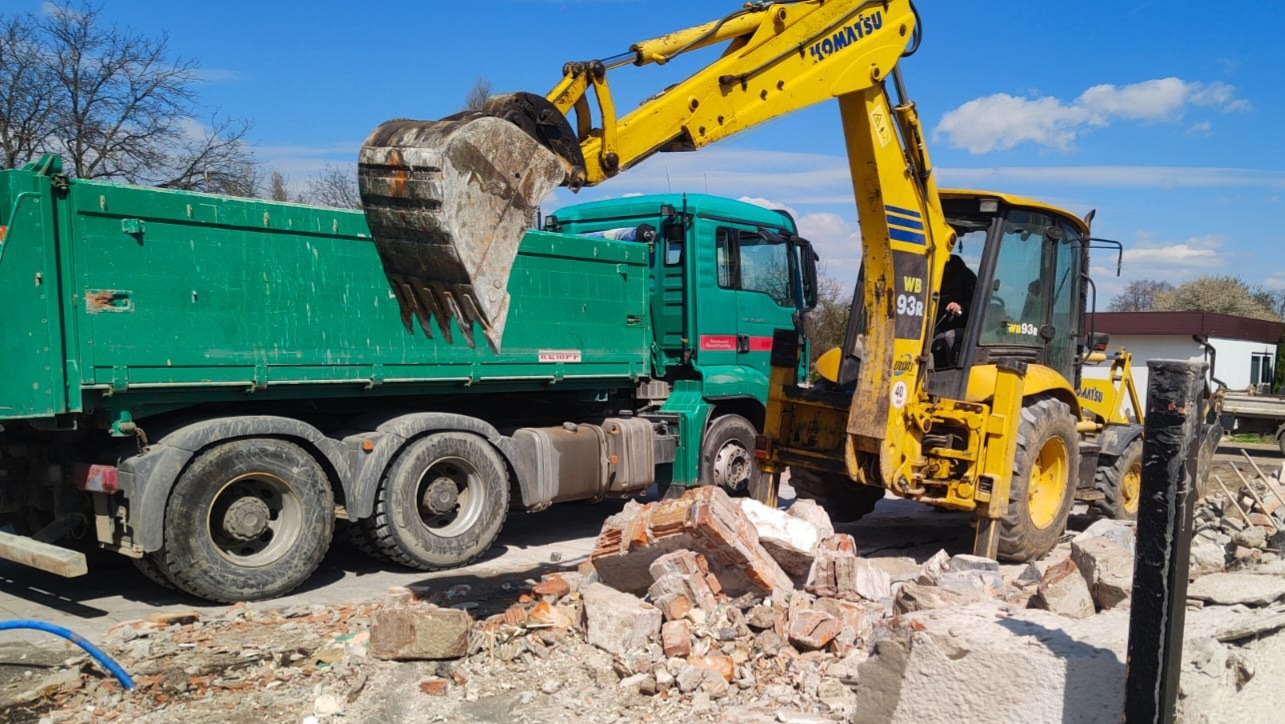 Żółta koparko-ładowarka Komatsu załadowuje gruz do zielonej ciężarówki MAN na tle błękitnego nieba. Widoczny fragment budynku w tle. Prace rozbiórkowe w toku.