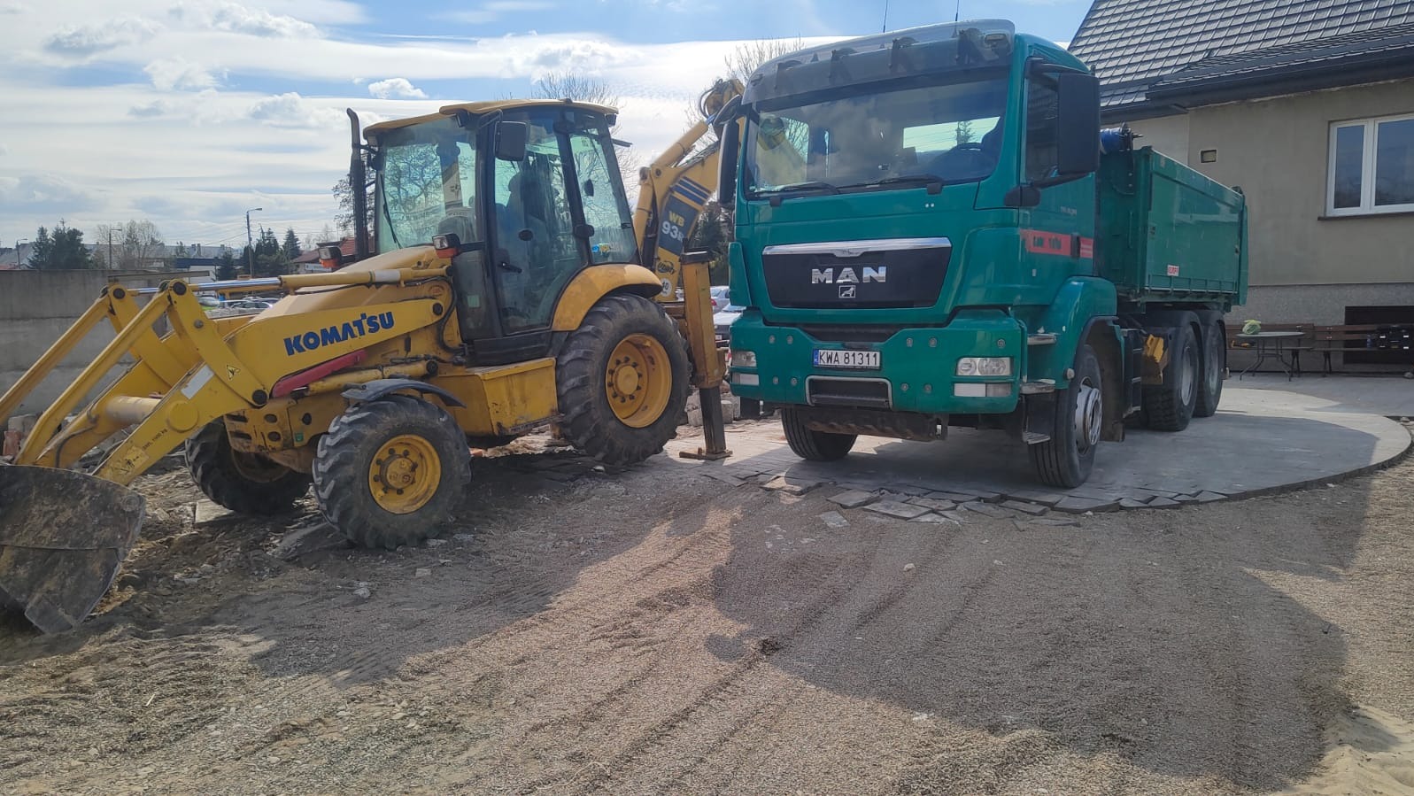 Żółta koparko-ładowarka Komatsu i zielona ciężarówka MAN na placu budowy z kostką brukową i piaskiem. Słoneczny dzień, błękitne niebo z chmurami.