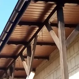 Drewniana altana przylegająca do murowanej ściany budynku, z widocznym orynnowaniem i drewnianą konstrukcją wsporczą.