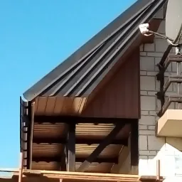 Fragment domu w trakcie prac dekarskich, widoczny nowy, ciemny dach z blachy trapezowej, elementy drewniane, rusztowanie i antena satelitarna na tle błękitnego nieba.