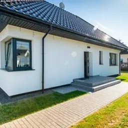 Biały, parterowy dom z ciemnym dachem i szarą kostką brukową prowadzącą do wejścia. Widoczne czarne rynny i okna z ciemnymi ramami. Świeżo położona, gładka elewacja.