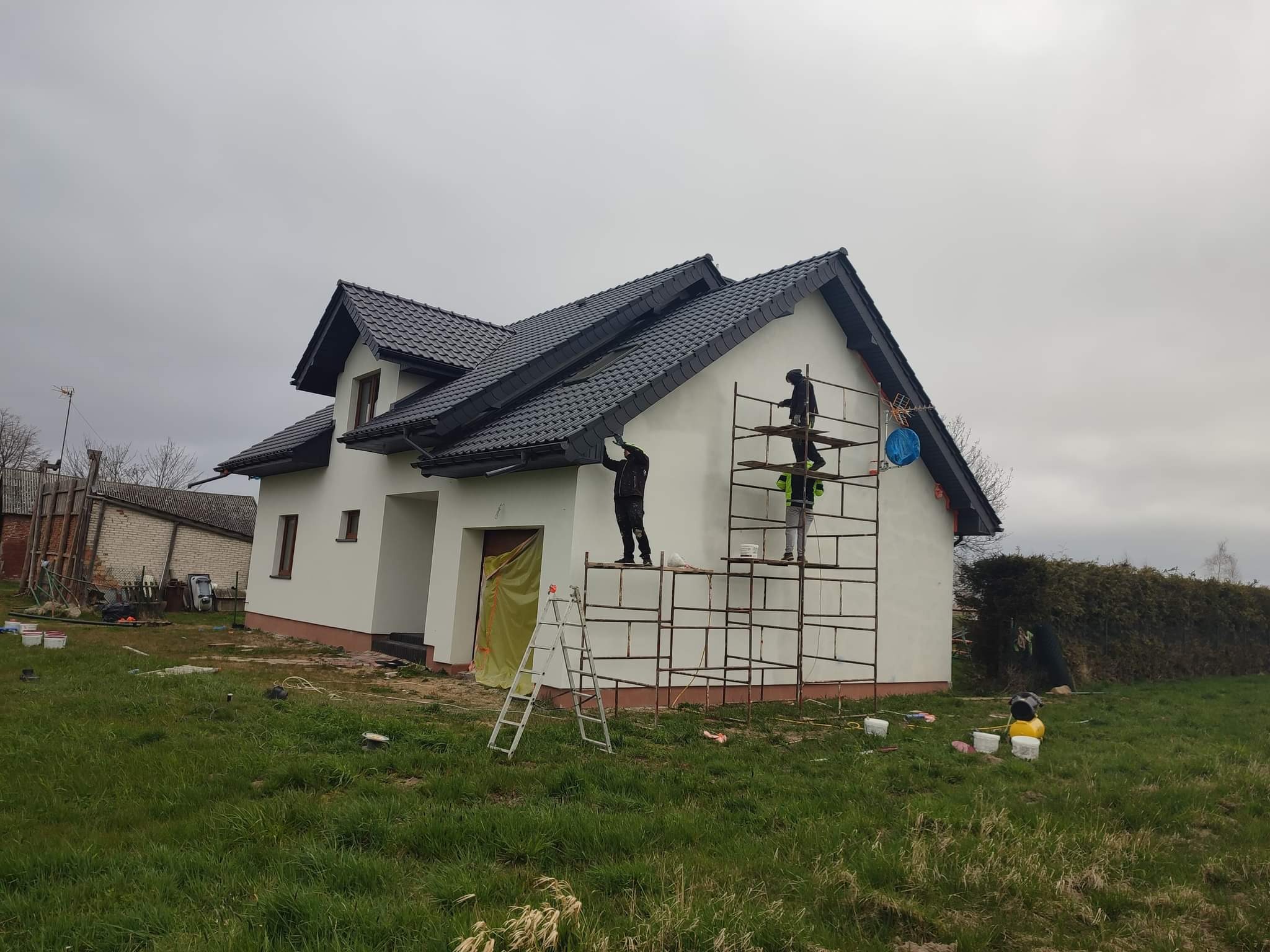 Malowanie elewacji domu jednorodzinnego z użyciem rusztowania i drabiny, widoczne otwarte puszki z farbą na trawie, pochmurne niebo.