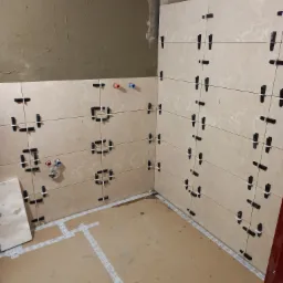 Ściany łazienki w trakcie układania płytek, widoczne krzyżyki dystansowe i przewody instalacji wodnej na tle surowego tynku.