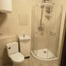 Jasna łazienka z kabiną prysznicową narożną, toaletą marki Cersanit, umywalką z szafką i grzejnikiem elektrycznym na ścianie wyłożonej płytkami imitującymi marmur.