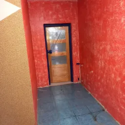 Ściana w kolorze czerwonym o nierównej fakturze w Charsznicy, drzwi zabezpieczone folią, niebieskie płytki podłogowe.