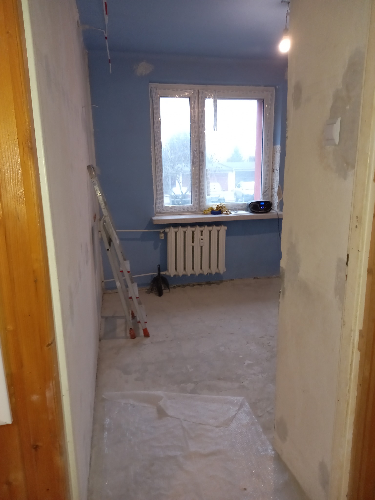 Pokój w trakcie remontu: ściany przygotowane do dalszych prac, zabezpieczona podłoga folią, drabina oparta o ścianę, widoczne okno z grzejnikiem i sprzętem grającym na parapecie.