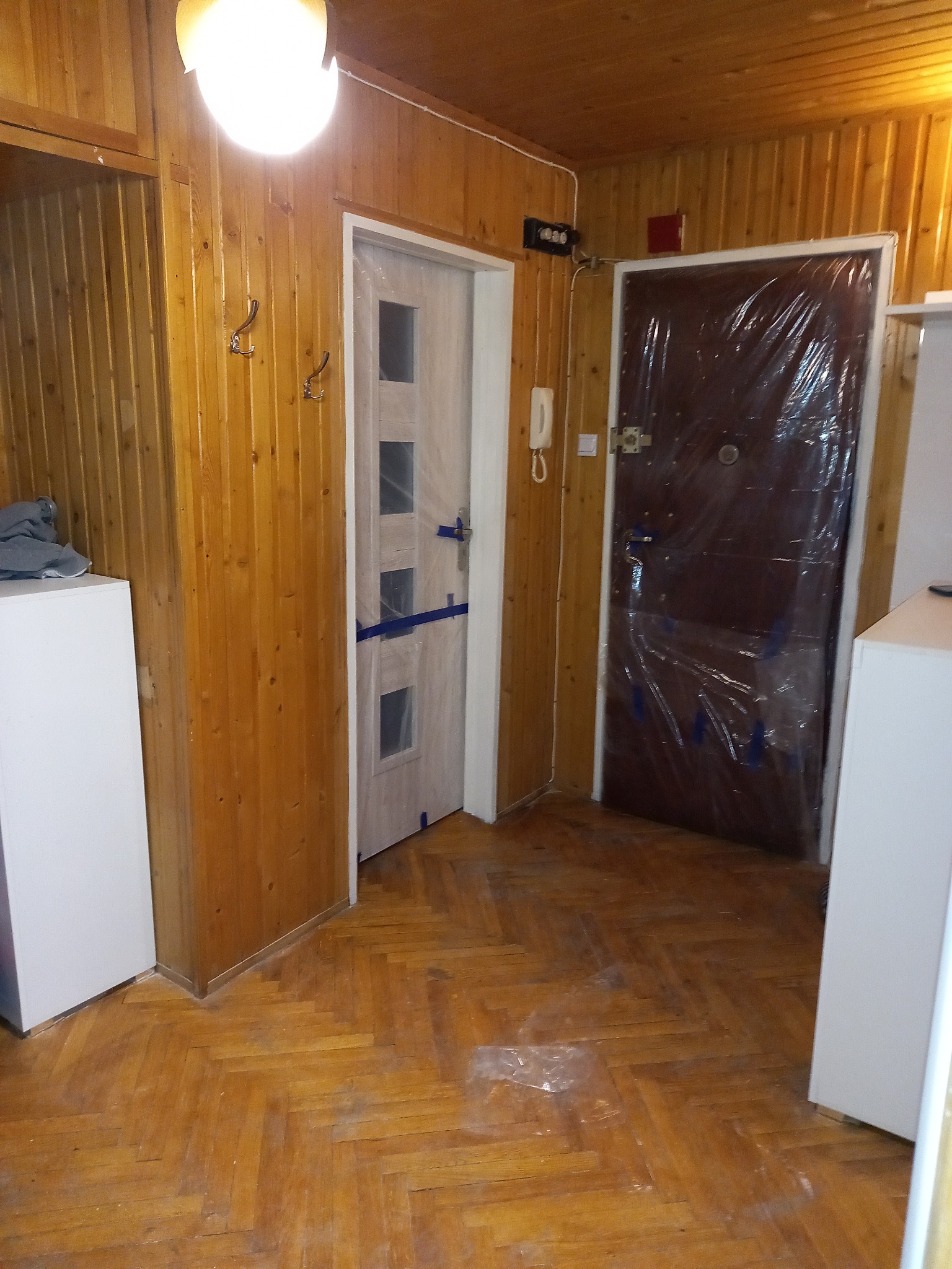 Dwie drzwi wewnętrzne, jedna jasna z okienkami, druga ciemna, obie zabezpieczone folią ochronną, widoczne na tle drewnianej boazerii ściennej i parkietu ułożonego w jodełkę.