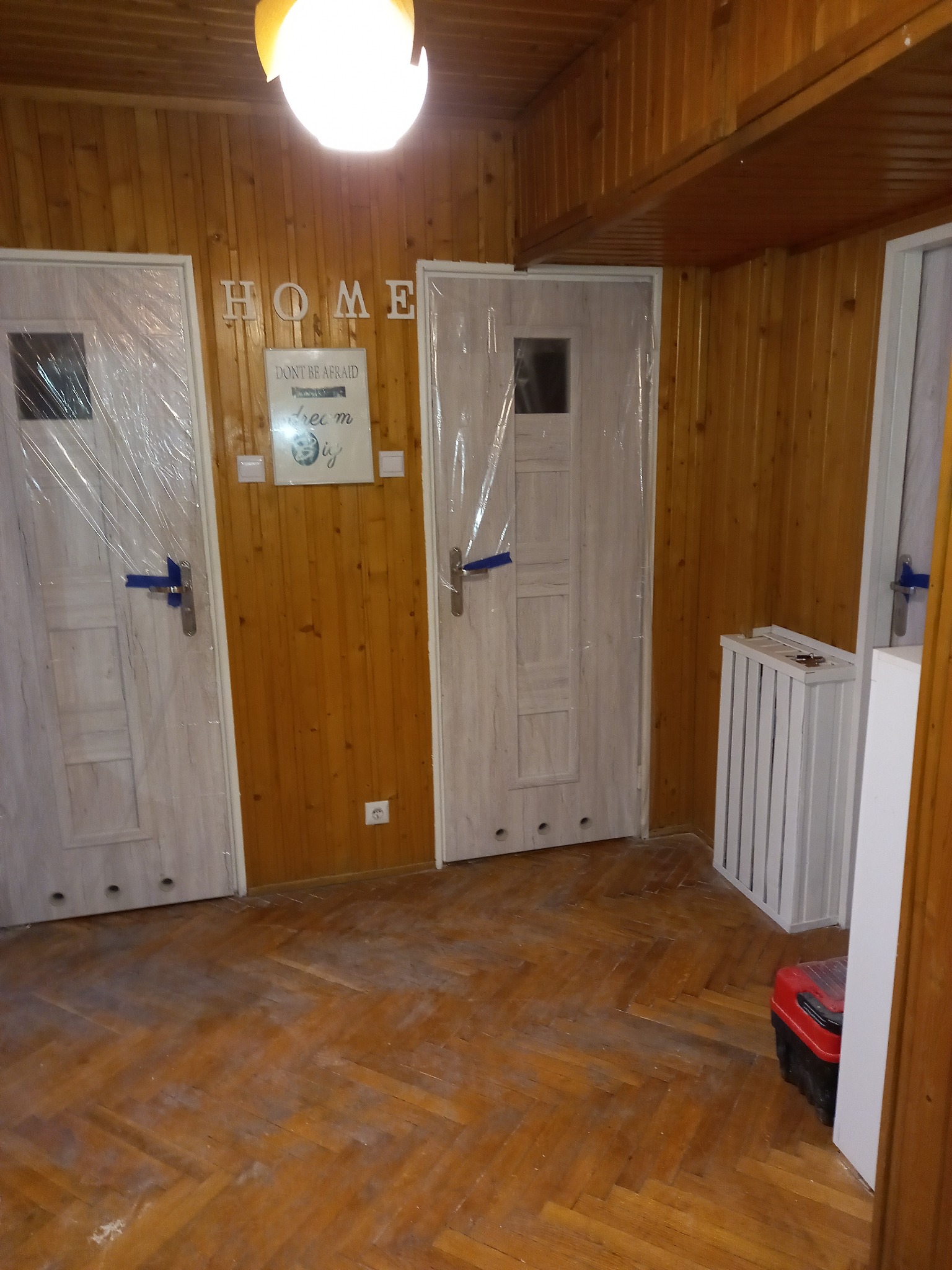 Pokój z drewnianą boazerią, dwiema nowymi, zabezpieczonymi folią drzwiami, drewnianą podłogą parkietową i białym grzejnikiem żeberkowym; widoczna skrzynka narzędziowa.