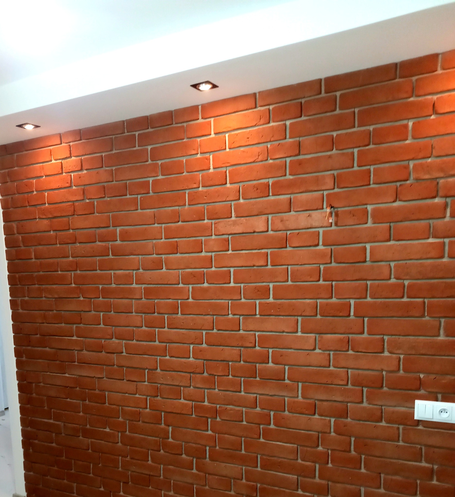 Ceglana ściana w odcieniach czerwieni z białymi fugami, oświetlona punktowymi lampami sufitowymi, widoczna w Charsznicy, z włącznikiem światła po prawej stronie.
