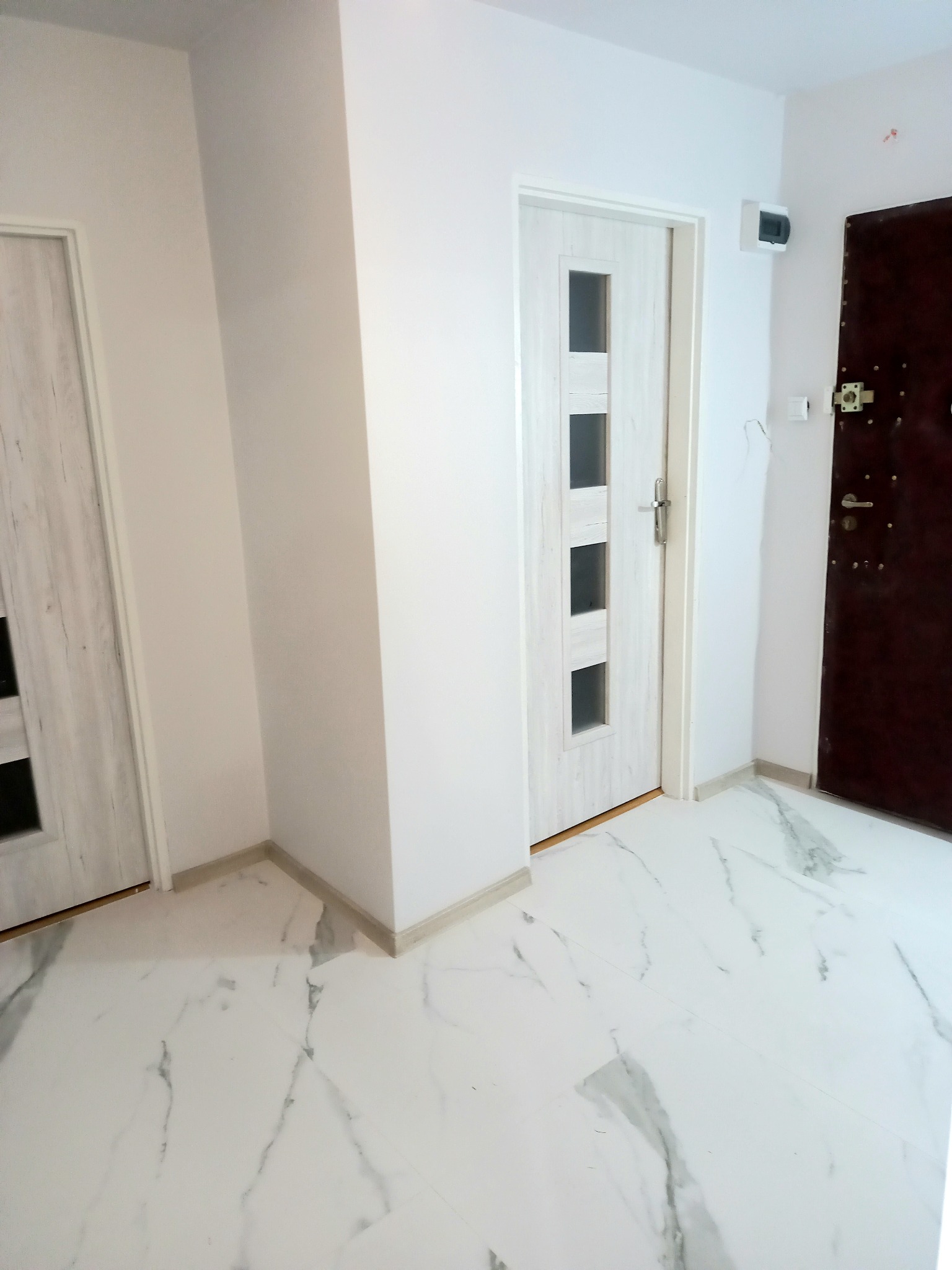 Wnętrze mieszkania po remoncie: białe ściany, jasna podłoga imitująca marmur, dwoje drzwi wewnętrznych z jasnego drewna oraz ciemne drzwi wejściowe.