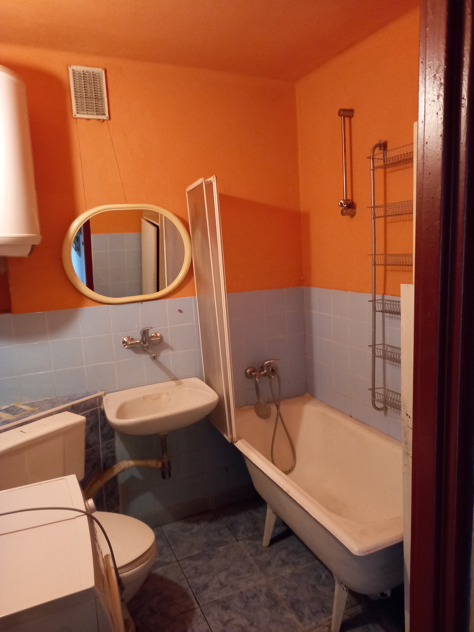 Pomarańczowo-niebieska łazienka z wanną na nóżkach, umywalką, toaletą i grzejnikiem elektrycznym, widok zza futryny drzwi.