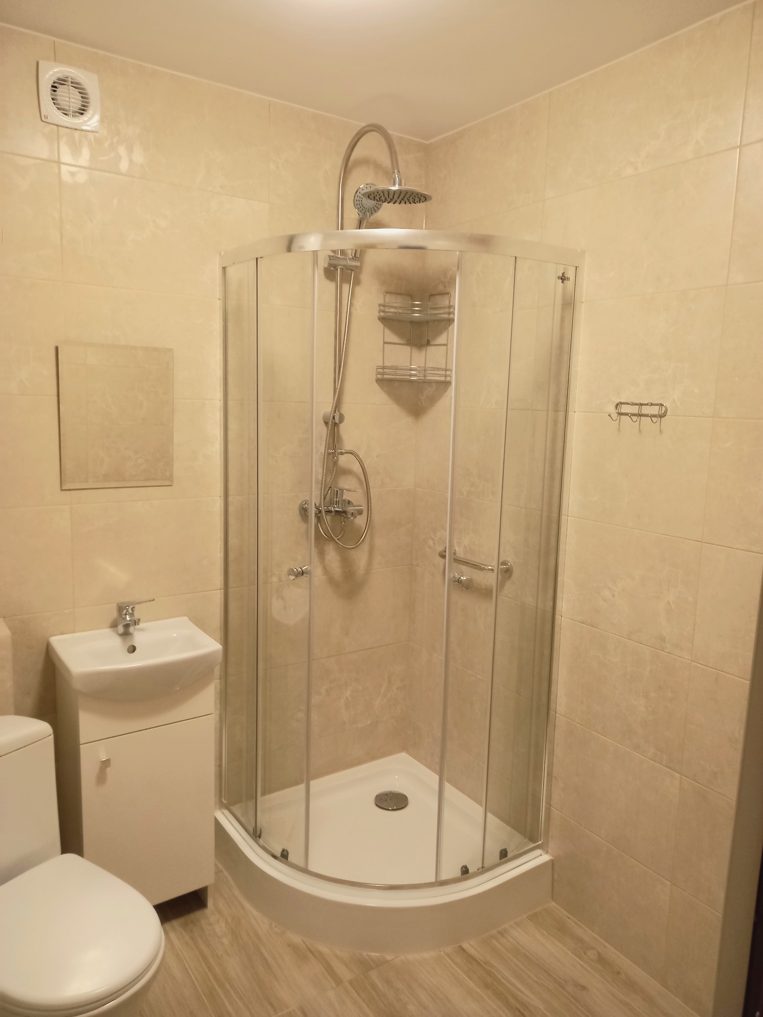 Jasna łazienka z narożną kabiną prysznicową ze szklanymi drzwiami, umywalką z szafką i wiszącym lustrem, wykończona beżowymi płytkami ceramicznymi.