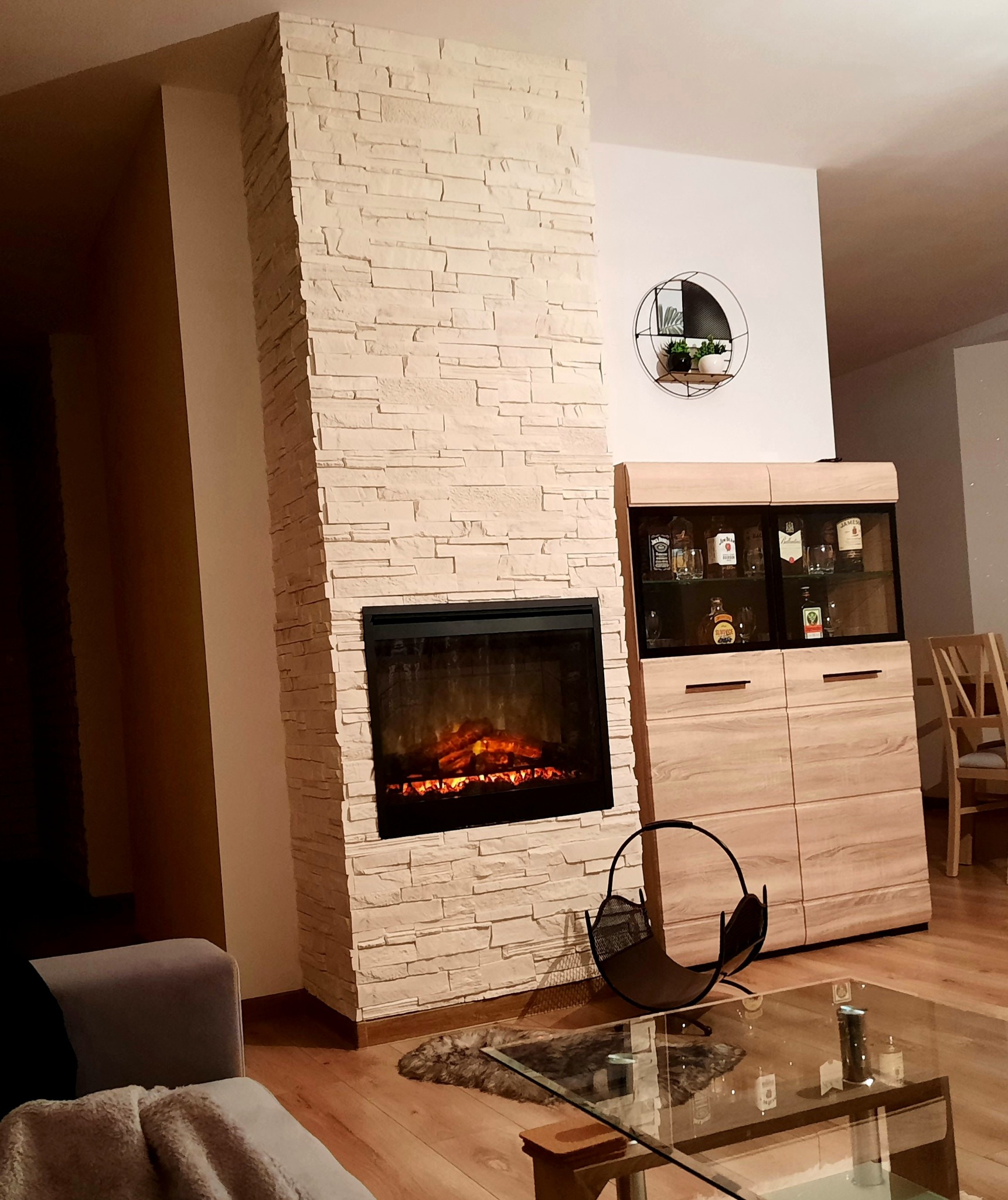 Salon z kominkiem obłożonym dekoracyjną, jasną cegłą, obok drewniana komoda z przeszkloną witryną, na ścianie okrągła półka z roślinami, na podłodze parkiet.