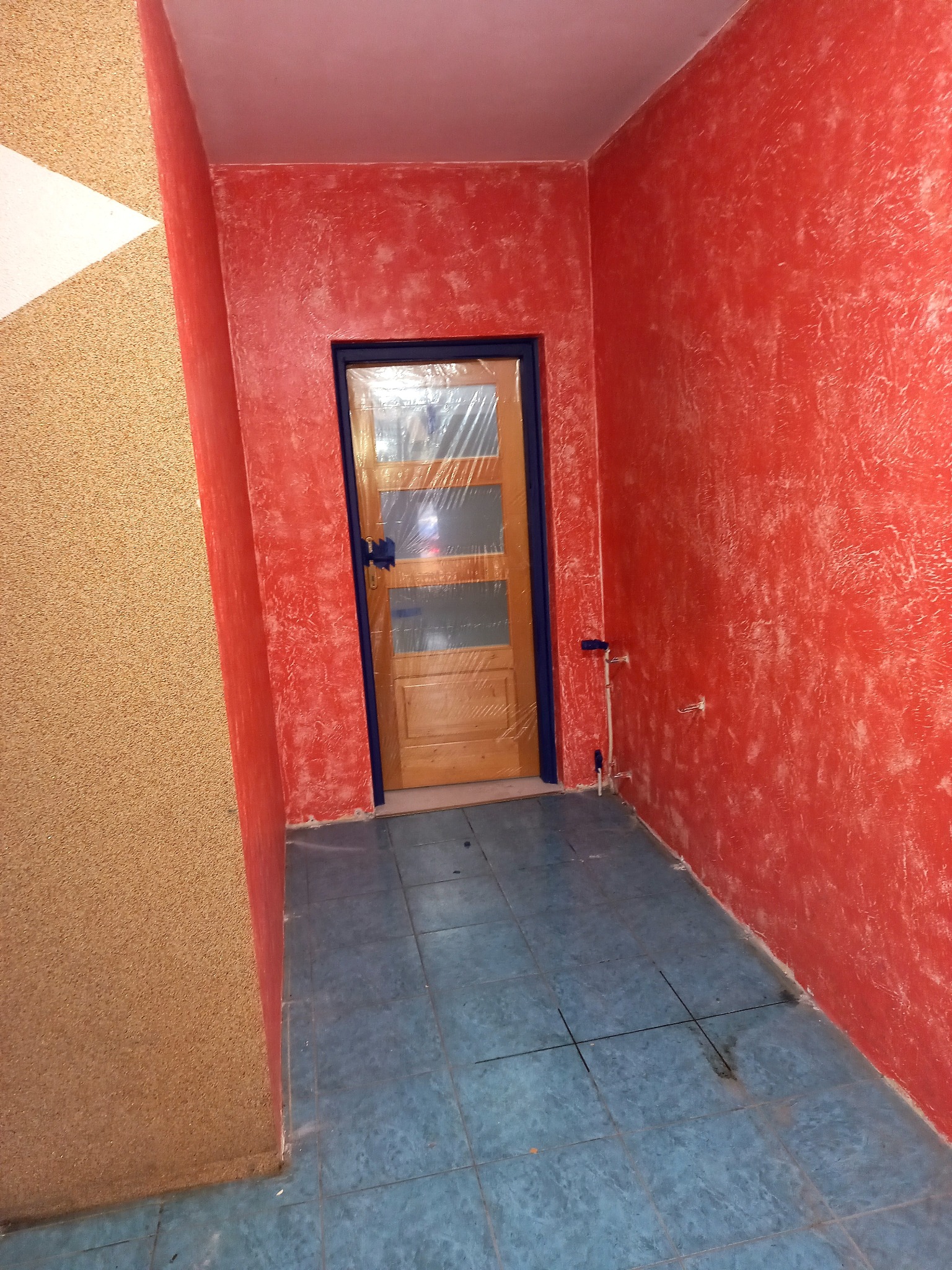 Ściana w kolorze czerwonym o nierównej fakturze w Charsznicy, drzwi zabezpieczone folią, niebieskie płytki podłogowe.