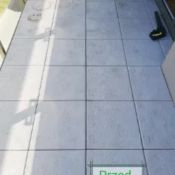 Szare płytki balkonowe przed czyszczeniem, widoczne zabrudzenia i cień barierki, dysza myjki ciśnieniowej Karcher w rogu.