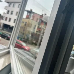 Otwarty biały plastikowy profil okienny, widok na budynki mieszkalne i zaparkowane samochody w pochmurny dzień.