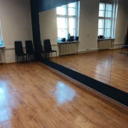 Sala z drewnianą podłogą, dużym lustrem na ścianie, trzema krzesłami pod ścianą i oknami z grzejnikami. Podłoga błyszczy po umyciu.