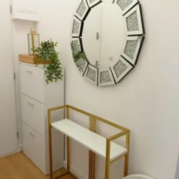 Nowoczesny przedpokój z lustrem o geometrycznym kształcie, białą szafką na buty, złotym stolikiem konsolą i zieloną rośliną doniczkową.