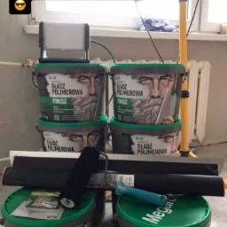 Kilka wiader z gładzią polimerową ułożonych w stos, obok wałki malarskie i szpachelki, w tle lampa budowlana i surowa ściana.