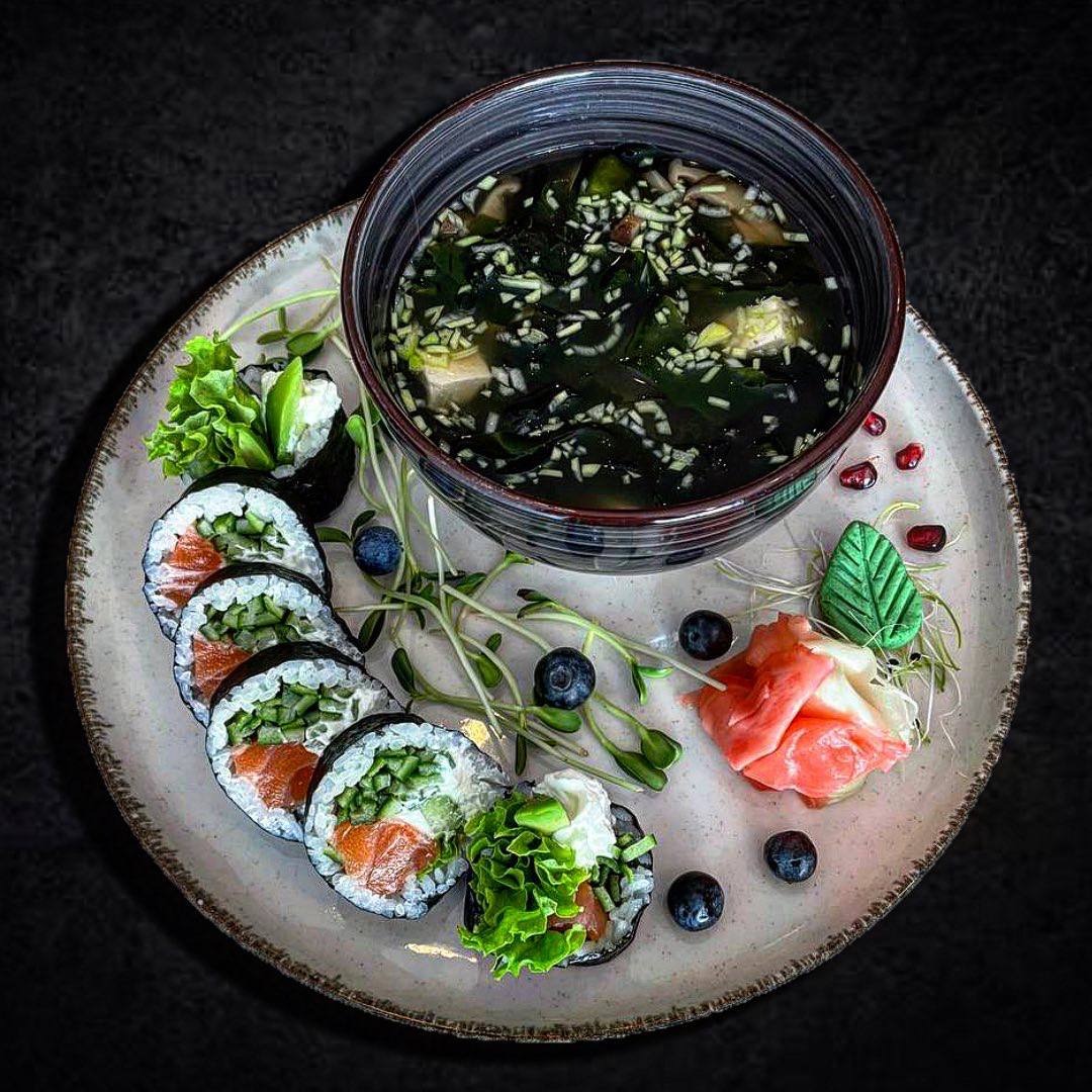 Elegancko podane danie: rolki sushi z łososiem i ogórkiem, zupa miso z tofu i wakame w ciemnej misce, marynowany imbir, borówki i kiełki na jasnym talerzu, ciemne tło.