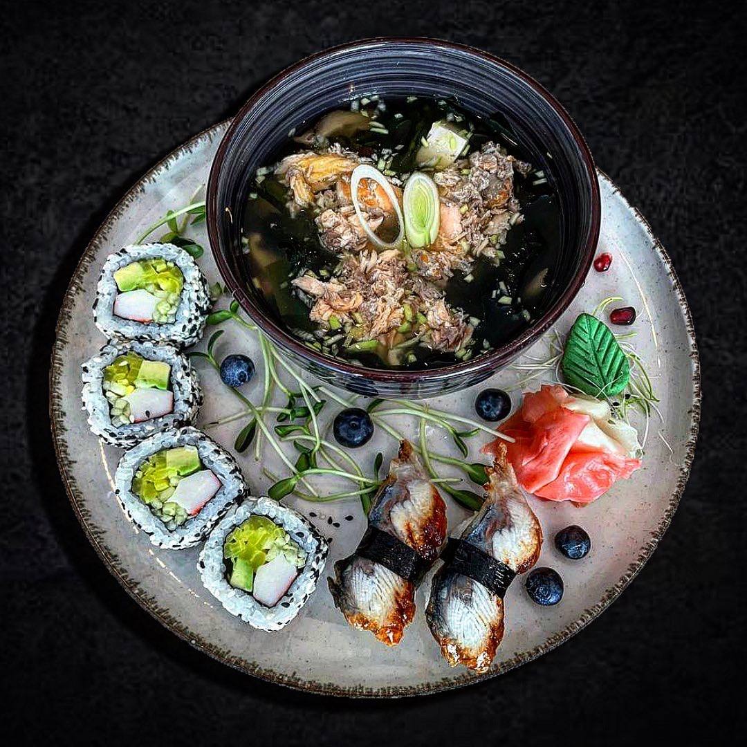 Zestaw sushi i zupy miso na szarej, zdobionej talerzu, z akcentami świeżych jagód i mikrozieleni. Widoczne rolki California z paluszkiem krabowym, nigiri z węgorzem oraz zupa miso z łososiem, tofu...
