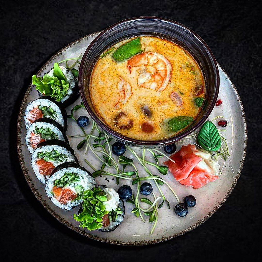 Tom yum z krewetkami w ciemnej misce, obok rolki sushi z łososiem i ogórkiem, udekorowane borówkami, pędami i marynowanym imbirem na ceramicznym talerzu z ciemnym tłem.
