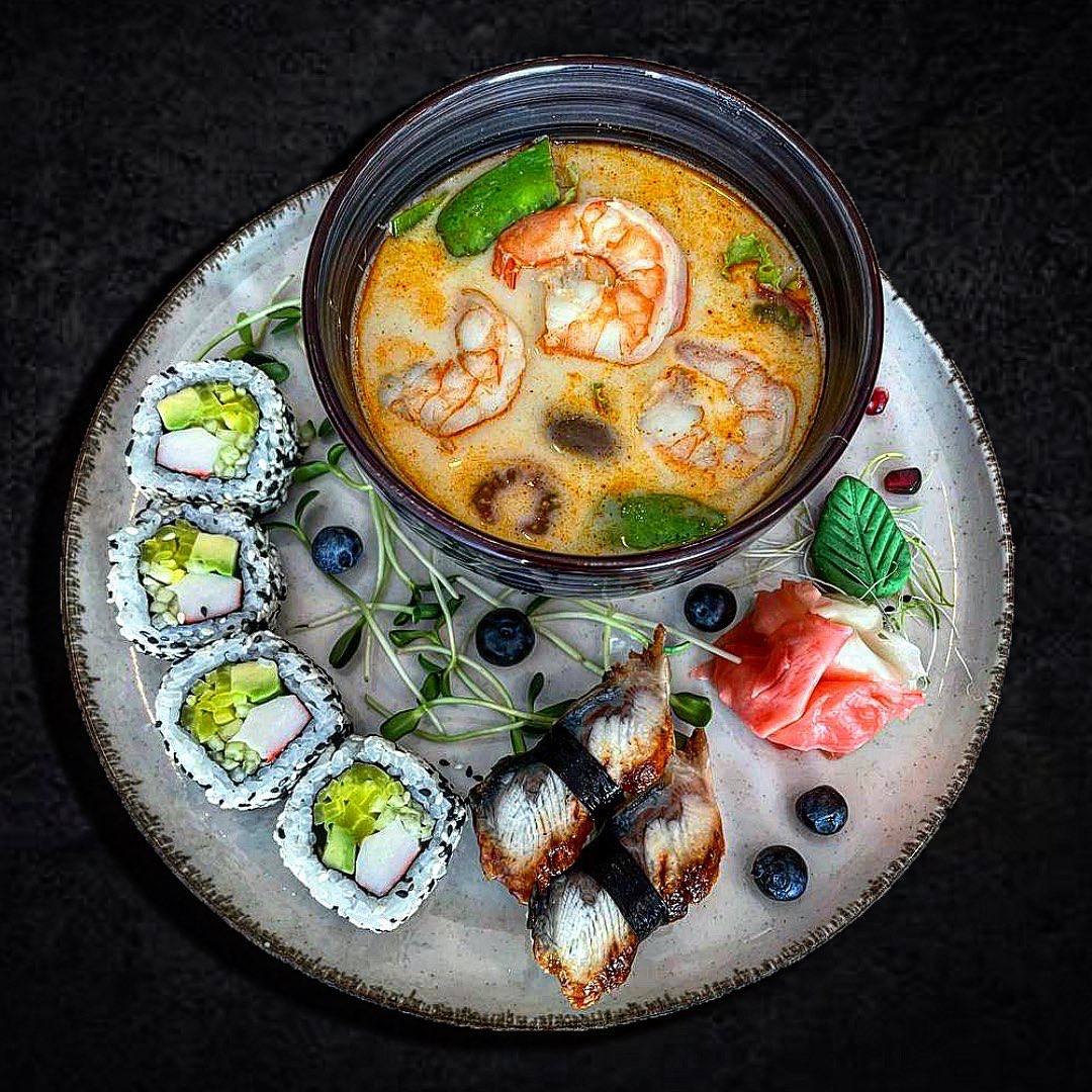 Tom yum z krewetkami w ciemnej misce obok sushi z paluszkiem krabowym i węgorzem na talerzu z szarymi brzegami, dekorowane borówkami i kiełkami.