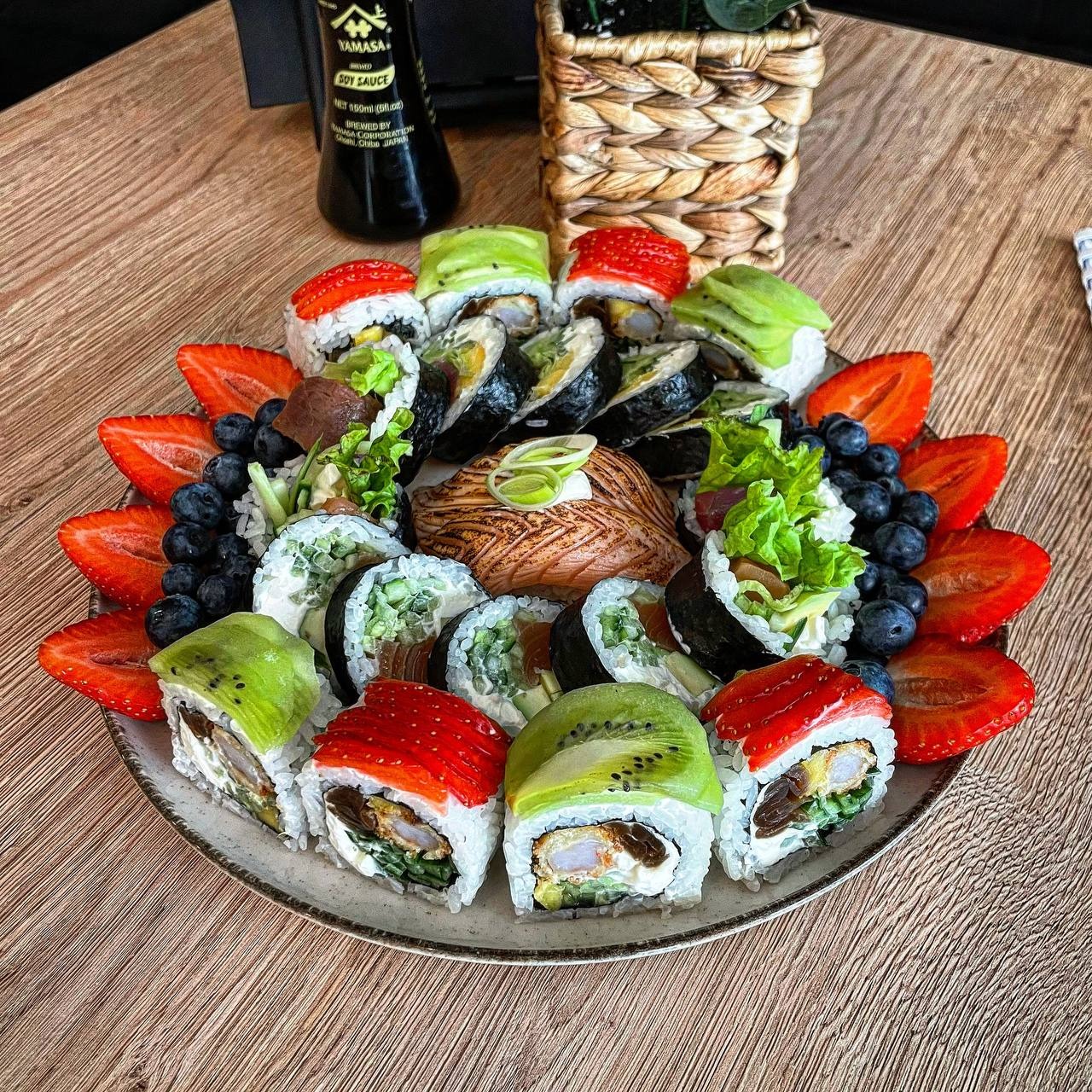 Wykwintna kompozycja sushi z owocami: rolki z kiwi, truskawkami, łososiem i sałatą, otoczone świeżymi truskawkami i borówkami, podane na drewnianym stole obok butelki sosu sojowego i plecionego...