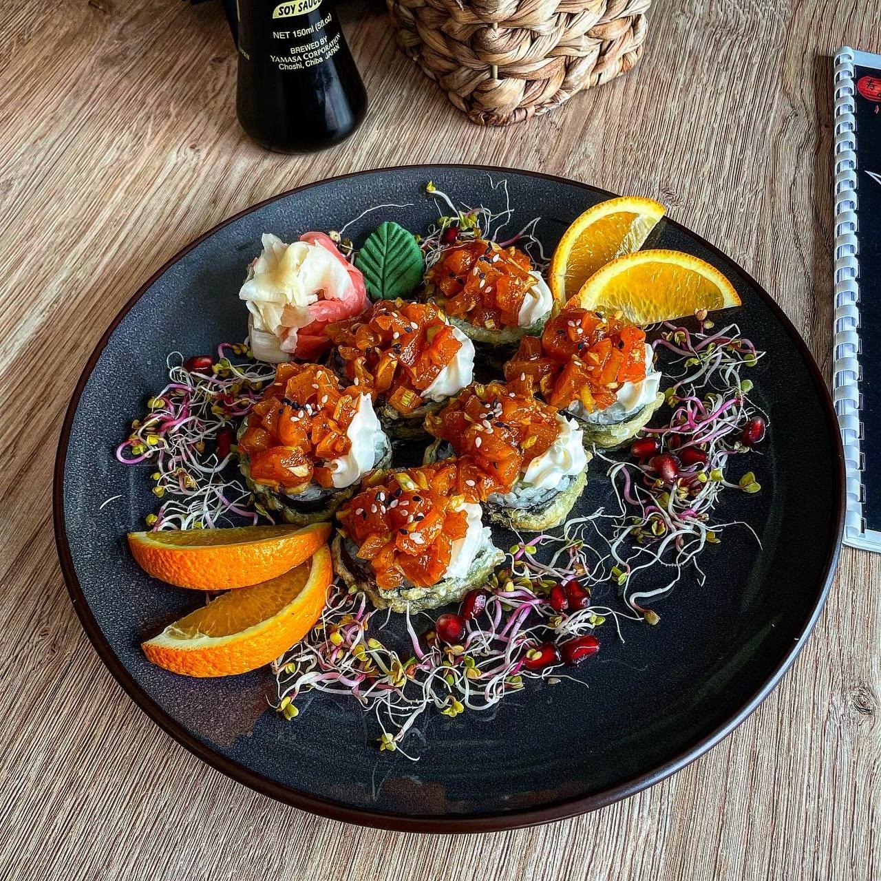 Czarny talerz z sześcioma rolkami sushi z tatarem z łososia, kremowym serem, ogórkiem i awokado, udekorowany kiełkami, granatem i plasterkami pomarańczy, obok butelka sosu sojowego i notes.